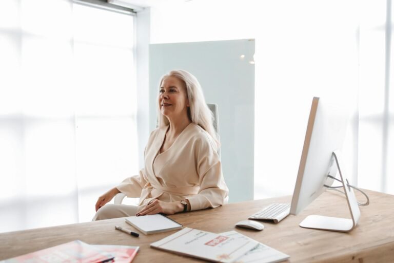Happy professional woman sitting at a desk with a computer and notes, conveying confidence in a bright office.